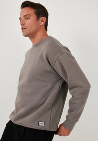 Grijze sweatshirt van zacht materiaal met een ronde hals en ribgebreide manchetten. Heeft een klein merklabel aan de onderkant van de linkerkant.