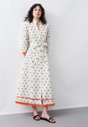 Žena s kučeravými vlasmi v bielych šatách s dlhým rukávom, drobnými kvetinovými vzormi, oranžovým lemom, predným rozparkom a opaskom viazaným v páse.