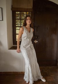 Gilet grigio in lino con scollo a V e dettagli con bottoni, abbinato a una gonna bianca in pizzo con orlo arricciato. Scarpe bianche semplici completano l'outfit.