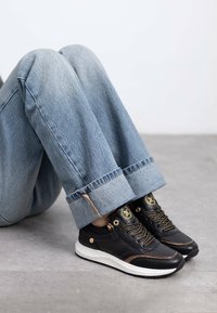 Zapatillas de cuero negro con acentos dorados y detalles en marrón; suela blanca. Pantalones de mezclilla azules con puños enrollados que exponen los zapatos.