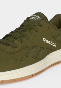 Αθλητικό παπούτσι Reebok σε ελιά πράσινο χρώμα με ταιριαστά κορδόνια, λευκή μεσαία σόλα, εξωτερική σόλα από καουτσούκ χρώματος καραμέλας και λογότυπα Reebok στη γλώσσα και στο πλάι.