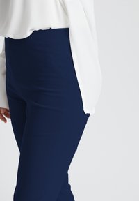 Donkerblauwe broek met een gladde textuur, hoge taille en slim fit, gecombineerd met een witte blouse met lange mouwen die over de broek valt.