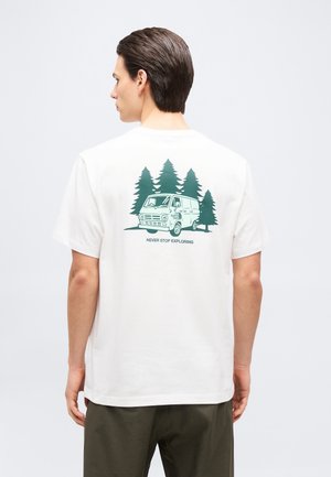 Hombre con cabello corto y oscuro que lleva una camiseta blanca con una furgoneta verde, pinos y el texto "Nunca dejes de explorar" en la parte trasera.