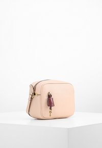 Kompaktní kožená taška v jemné broskvové barvě, vybavená zipovým zapínáním, obdélníkovým tvarem a bordo akcentem na štítku s ozdobou ve tvaru klíče.