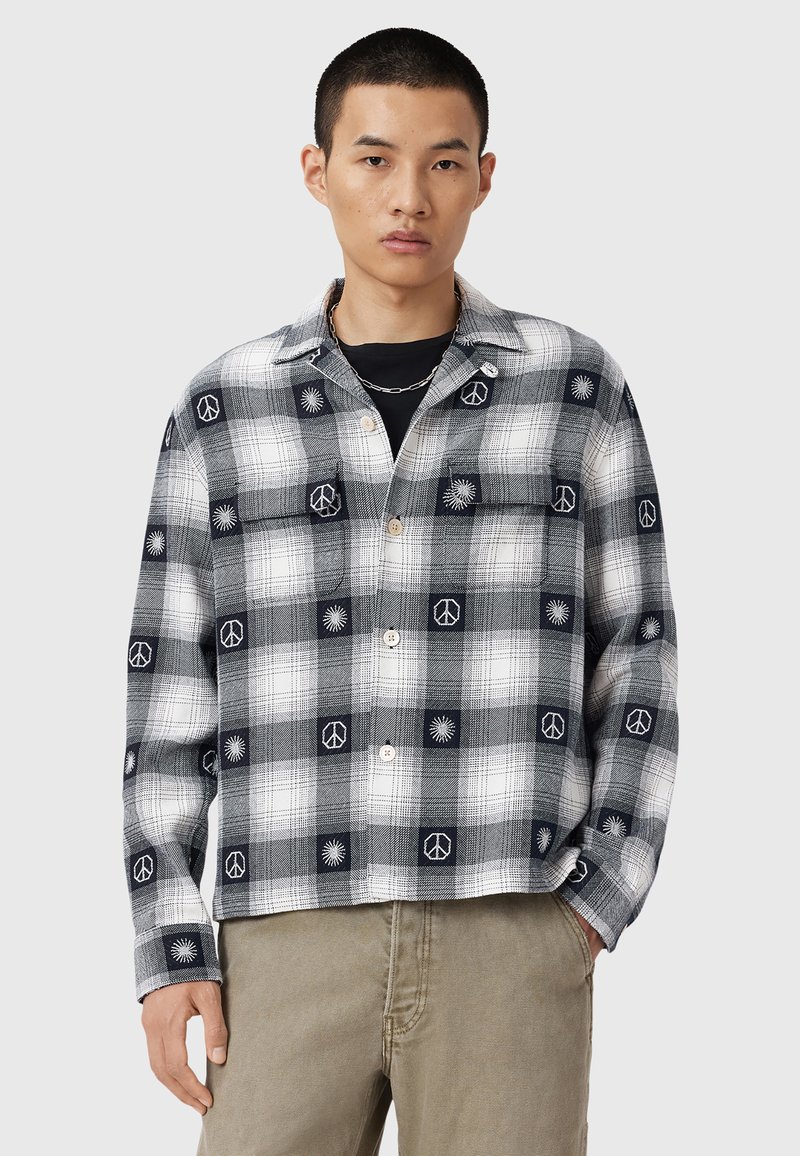 Chemise à carreaux noir et blanc avec des motifs brodés de signes de paix et de soleil, à coupe décontractée et à fermeture par boutons. Tissu en coton épais.