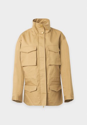 Person trägt eine beige Jacke mit mehreren Taschen, ein weißes Hemd mit Kragen, schwarze weite Lederhosen und schwarze Loafers auf einfarbigem Hintergrund.