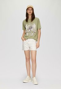 Oliwkowa bawełniana koszulka z nadrukiem, zestawiona z kremowymi szortami dżinsowymi i jasnymi sneakersami. Prosta czapka dopełnia całość.