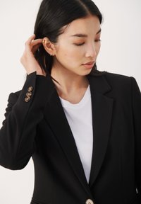 Černý blazer s klopami, který má zlaté knoflíky na rukávech. Nosený přes bílé tričko, hladká textura a dobře padnoucí design.