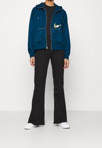 Person klädd i en blå dragkedjetröja med en gul Nike-logga, svarta byxor och vita sneakers, stående mot en enkel vit bakgrund.