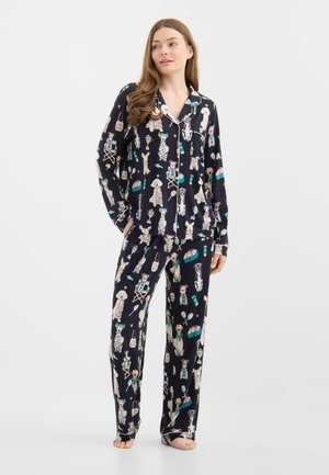 Donna che indossa un pigiama nero con motivi colorati di cani e strumenti per la toelettatura, in piedi a piedi nudi con le mani nelle tasche, che guarda leggermente a destra.