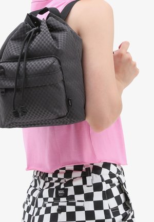 Zaino con coulisse a quadri neri, tessuto testurizzato, tasca frontale con zip, indossato sopra una maglietta rosa. Pantaloni a quadri neri e bianchi visibili.