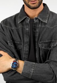 Orologio d'argento con quadrante blu e accenti rossi su cinturino in acciaio inox, indossato su un polso coperto da una giacca di jeans grigia con bottoni a pressione.