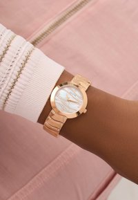 Reloj redondo de oro rosa con una esfera de nácar, diseño en forma de ola y acentos de cristal en la correa, llevado en una muñeca con una manga clara.