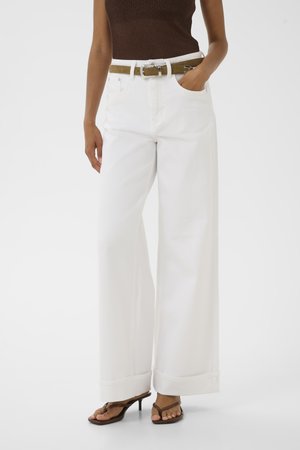 Persona que lleva pantalones blancos de pierna ancha con puños, una camiseta marrón sin mangas, cinturón verde oliva y sandalias marrones con tacón, de pie sobre un fondo blanco.