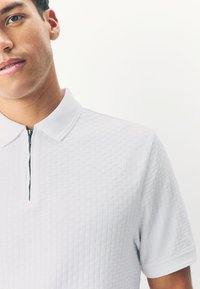 White polo shirt with a textured pattern, featuring a short zipper and a classic collar. Soft fabric with a subtle, checkered design.