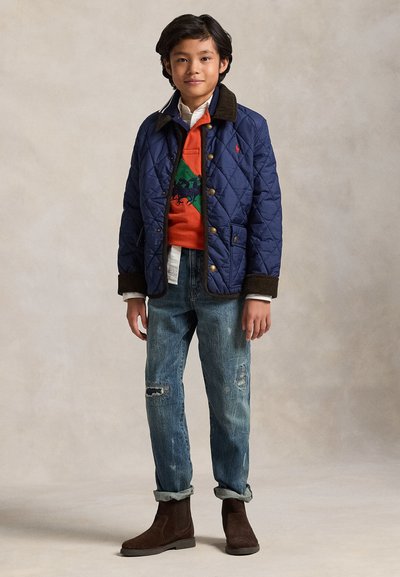 Garçon portant une veste matelassée bleu marine, un pull orange avec un motif cheval de polo, un jean bleu retroussé et des bottes marron, debout devant un fond uni.