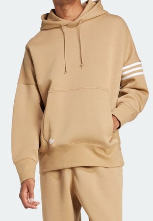 Hombre vestido con sudadera con capucha beige y pantalones deportivos a juego, la sudadera tiene bolsillo frontal, cordones y franjas blancas en la manga izquierda.