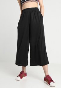 Culottes negras con pliegues y una cinturilla elástica, que cuentan con bolsillos laterales. Combinados con zapatillas rojas con acentos dorados.