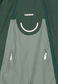 Macaco impermeável verde com um detalhe circular refletor, dois bolsos laterais com fecho de correr e um fecho de correr de comprimento total. Apresenta um acabamento texturizado.