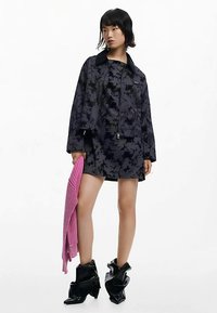 Musta- ja harmaakuvioinen kukkakuosinen takki, jossa on vetoketjusuljenta edessä, yhdistetty vastaavaan mekkoon. Pitelee vaaleanpunaista neulepaitaa; jalassa kiiltävät mustat nilkkurit.