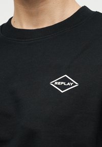 Μαύρο πουλόβερ με στρογγυλή λαιμόκοψη και μικρό λευκό λογότυπο διαμαντιού στο στήθος που εμφανίζει τη λέξη "REPLAY".