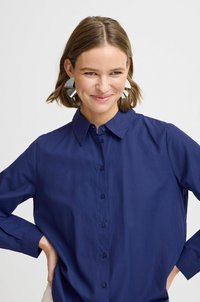 Navy blue button-up shirt with a collar and long sleeves, featuring a smooth texture and a relaxed fit. Large, silver geometric earrings.