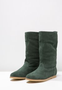 Une paire de bottes en daim vert foncé, mi-mollet, avec des semelles en caoutchouc beige plates sur une surface blanche, contre un fond clair uni.