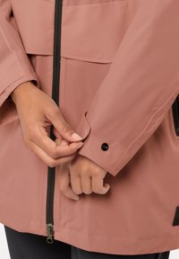Giacca mauve resistente all'acqua con zip frontale, polsini elastici e dettagli in bottone. Presenta una texture liscia e accenti neri.