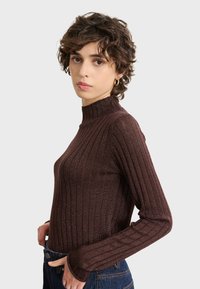 Brauner, gerippter Rollkragenpullover mit einer strukturierten, glänzenden Oberfläche und langen Ärmeln. Getragen mit hoch geschnittenen Denim-Jeans, die eine figurbetonte Silhouette aufweisen.