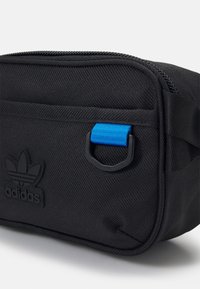 Kompakt svart Adidas-väska med framdragkedja, en blå remslinga och Adidas trefoil-logotyp prentad på det främre tyget.