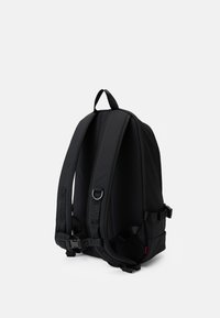 Sac à dos noir avec des bretelles réglables rembourrées, une poignée supérieure, des boucles latérales et une petite étiquette rouge sur fond blanc.