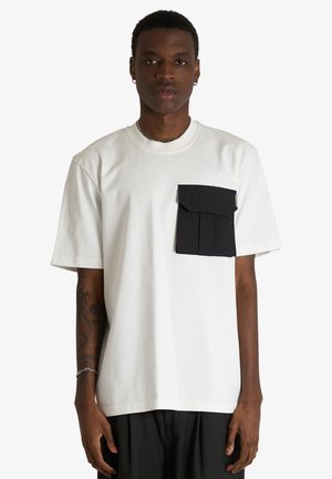 Giovane uomo che indossa una t-shirt bianca a maniche corte con una grande tasca nera sul petto sinistro, in piedi davanti a uno sfondo semplice.