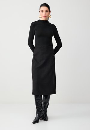 Kobieta ubrana w dopasowaną czarną bluzkę z długim rękawem, czarną spódnicę midi z bocznymi guzikami, czarne kozaki do kolan oraz złote kolczyki koła.