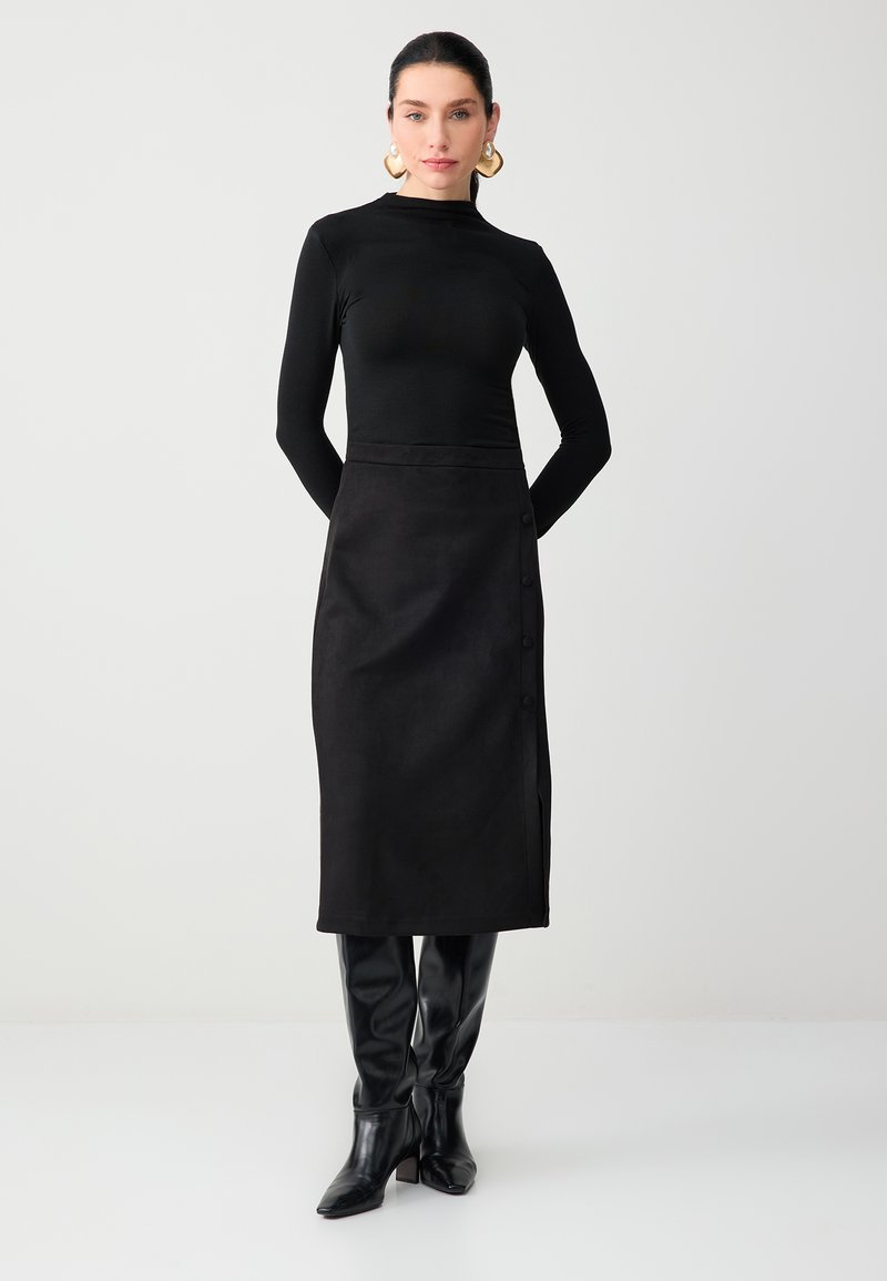 Femme portant un haut noir à manches longues ajusté, une jupe midi noire avec des boutons sur le côté, des bottes noires montantes jusqu'au genou et des boucles d'oreilles créoles dorées.