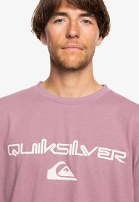 Svetloružové bavlnené tričko s okrúhlym výstrihom, s veľkým bielym logom "Quiksilver" na hrudi. Mäkkučký materiál, voľný strih.