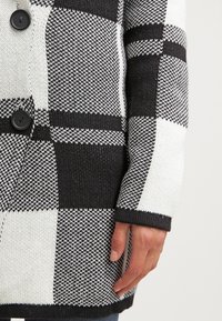 Cardigan en maille à carreaux noirs et blancs, doté de gros boutons noirs, de poignets côtelés et d'un motif texturé sur le tissu.