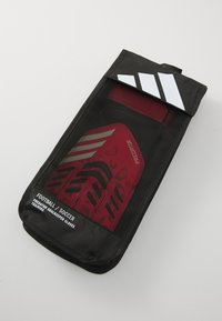 Sac en maille noir contenant des gants de gardien de but rouges avec des accents noirs. Il présente un branding blanc et un dessus plié avec un onglet pour la fermeture.