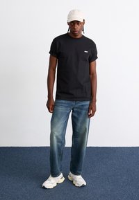 Zwarte katoenen t-shirt met korte mouwen en een klein wit logo, gecombineerd met vervaagde blauwe straight-leg jeans en witte sneakers. Beigekleurige pet.