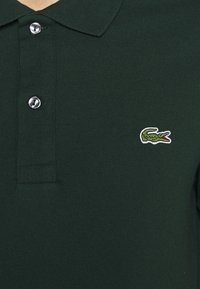 Tmavě zelené polo tričko vyrobené z texturované látky, s límečkem a zapínáním na tři knoflíky. Vyšívané logo krokodýla na levé hrudi.