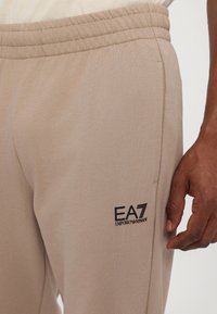 Beige sweatpants van zacht materiaal, met een elastische tailleband en zijzakken. Zwart EA7-logo nabij de heup. Gladde textuur.