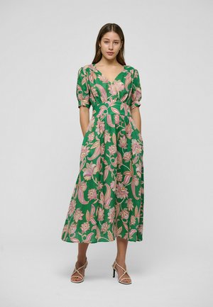 Woman wearing a green floral midi dress with puff sleeves and a V-neck, paired with beige strappy high-heeled sandals, standing against a gray background.