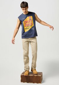 Mörkblå ärmlös skjorta med "CASEY JONES" grafisk design i rosa och gult, kombinerad med beige byxor och bruna stövlar, stående på en träbox.