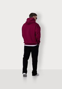 Bordeauxfarbener Hoodie mit einer großen Rückentasche, körpernahen Bündchen und einer Kapuze mit Kordelzug. Kombiniert mit schwarzen Hosen und weißen Schuhen, von hinten betrachtet.