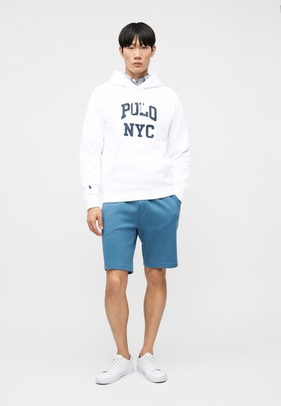 Άνδρας που φοράει άσπρο φούτερ Polo NYC, μπλε σορτς και άσπρα αθλητικά παπούτσια, στέκεται μπροστά σε απλό φωτεινό φόντο.