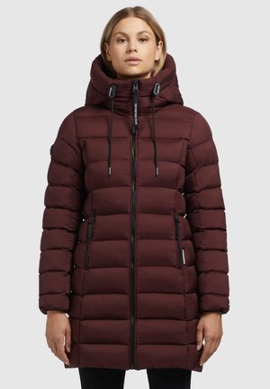 Femme portant un long manteau d'hiver matelassé bordeaux avec un col montant, une capuche ajustable et une fermeture éclair noire, se tenant devant un fond uni.