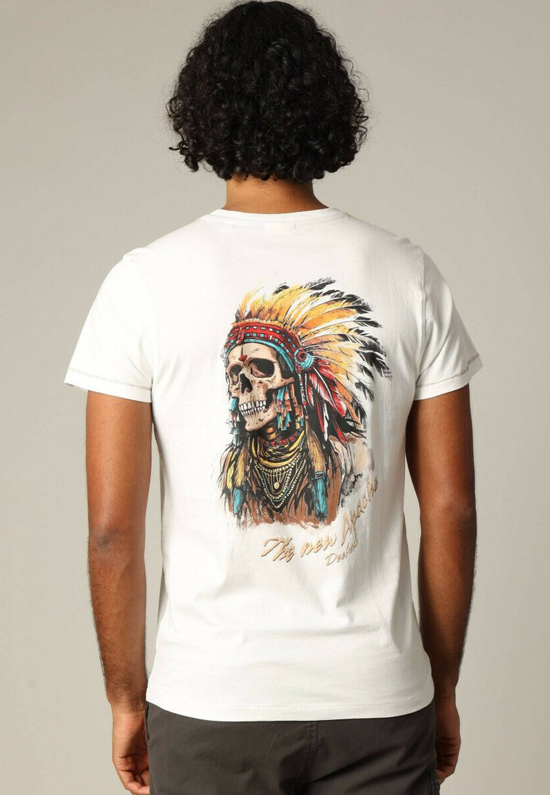 T-shirt en coton blanc avec un graphique d'un crâne orné d'un couvre-chef en plumes et de bijoux ; des accents colorés à travers le design.