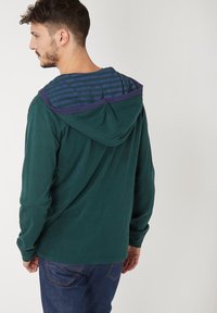 Sudadera verde de manga larga con una capucha interior a rayas en azul y púrpura. Presenta un corte relajado y está hecha de material de algodón suave.