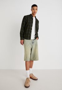 Green plaid button-up shirt over a white T-shirt, paired with light denim shorts and tan sneakers. Features white socks and subtle accents.