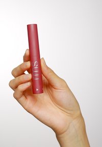Produit à lèvres cylindrique bordeaux avec un branding argenté, tenu dans une main, présentant une finition mate lisse et des accents triangulaires.