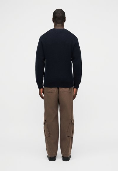 Άνδρας που στέκεται πλάτη με μαύρο μακρυμάνικο πουλόβερ, καφέ παντελόνι καρό και μαύρα παπούτσια μπροστά σε μια απλή ανοιχτόχρωμη επιφάνεια.